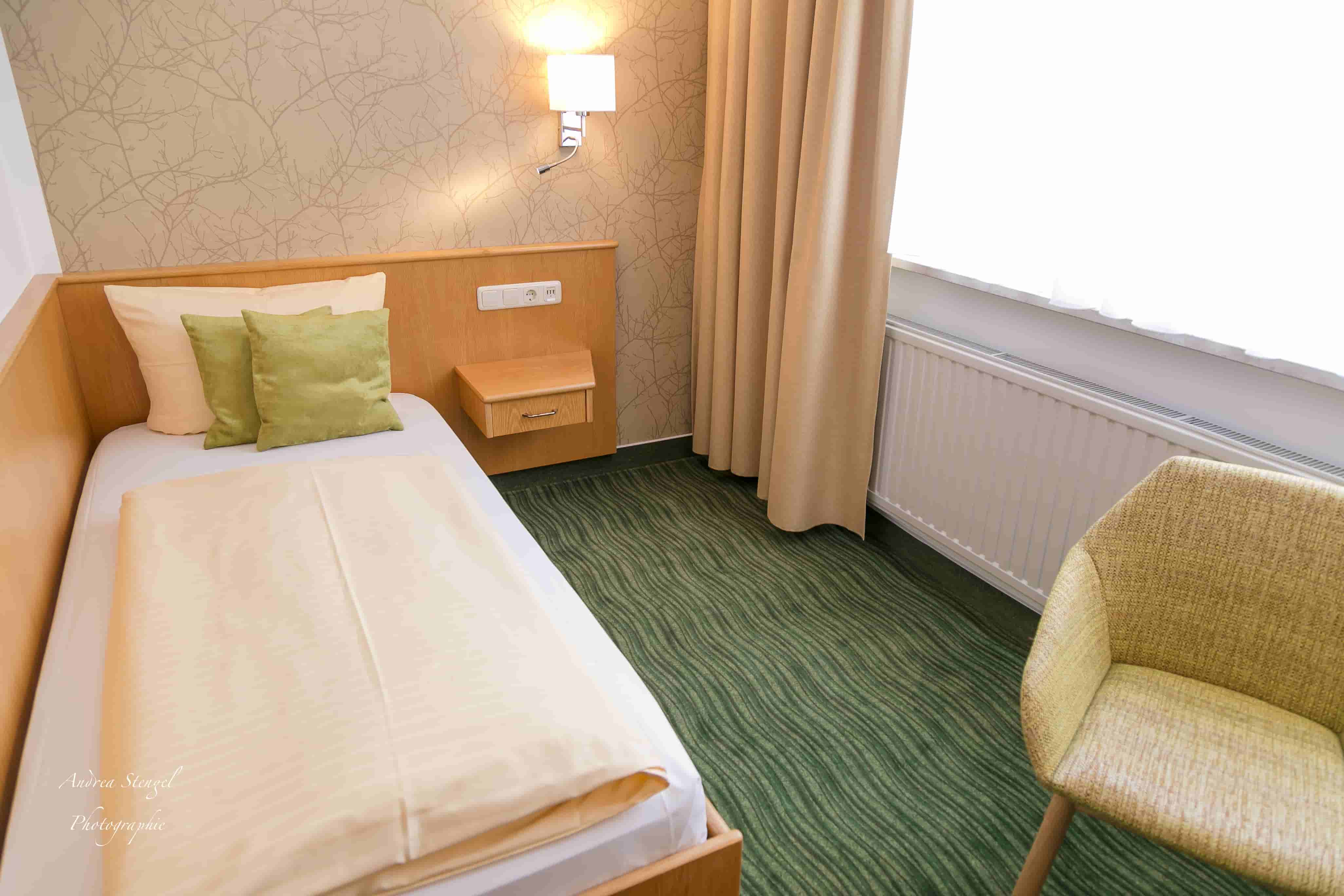 Einzelzimmer mit Bett, beigen Vorhängen, grünem Teppich und einem hellen Sessel.