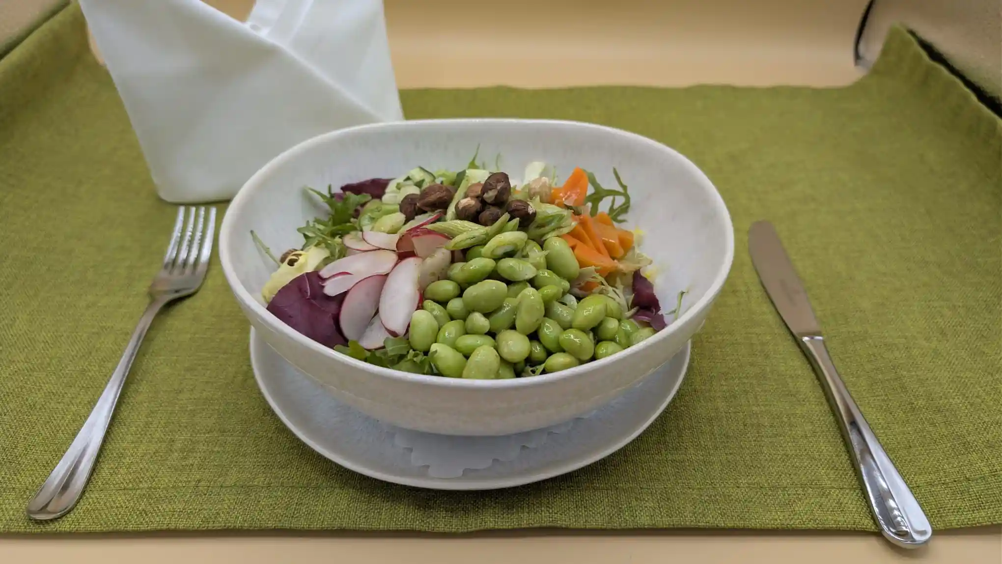 Schüssel mit frischem Salat, darunter Edamame, Radieschen, Karotten und Nüsse, auf grünem Tischset mit Messer und Gabel.