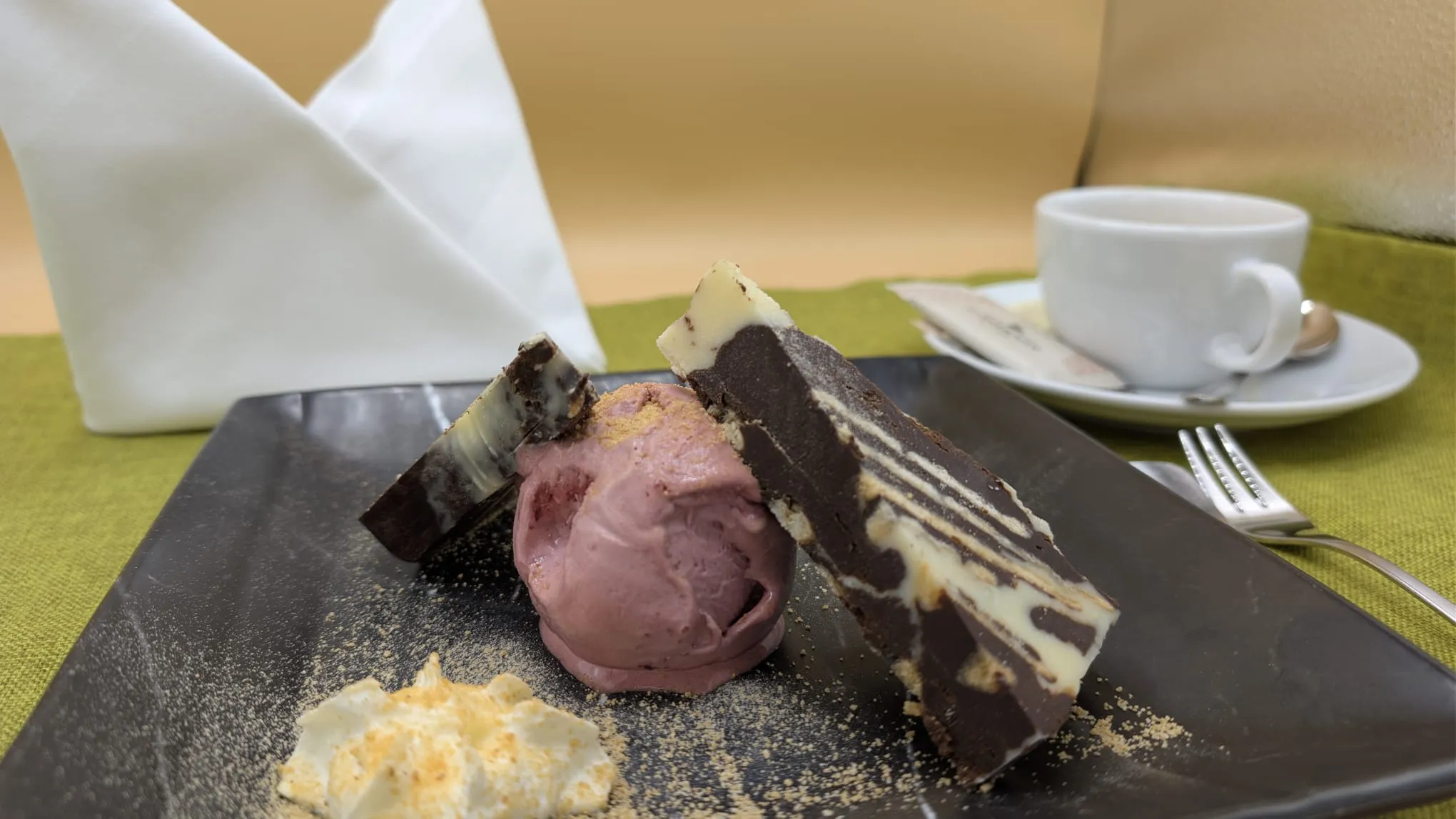 Dessert mit Schokoladenmousse, Himbeereis und Sahne auf schwarzem Teller mit Kaffee im Hintergrund.